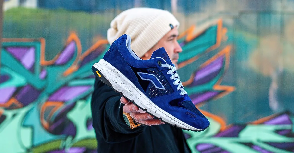 Hombre elegante presentando una zapatilla azul contra un vibrante graffiti urbano, exhibiendo moda moderna.