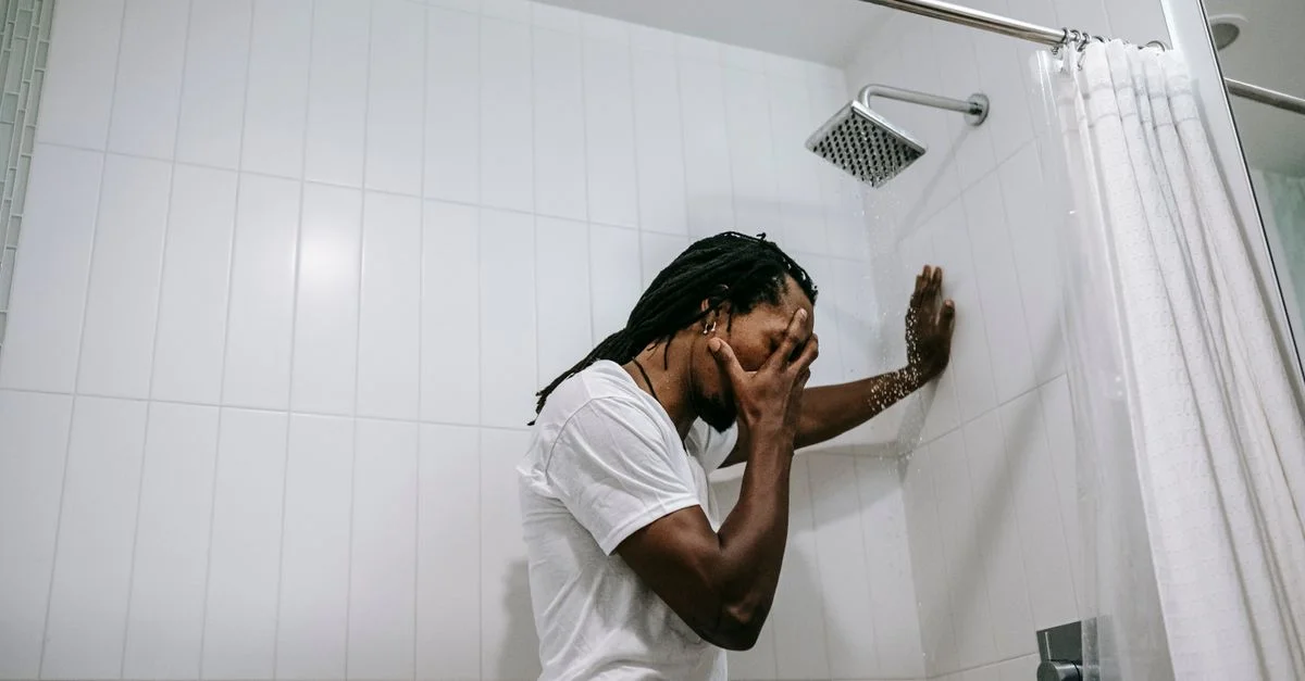 Un hombre adulto está en una ducha, visiblemente estresado y melancólico, cubriéndose la cara.