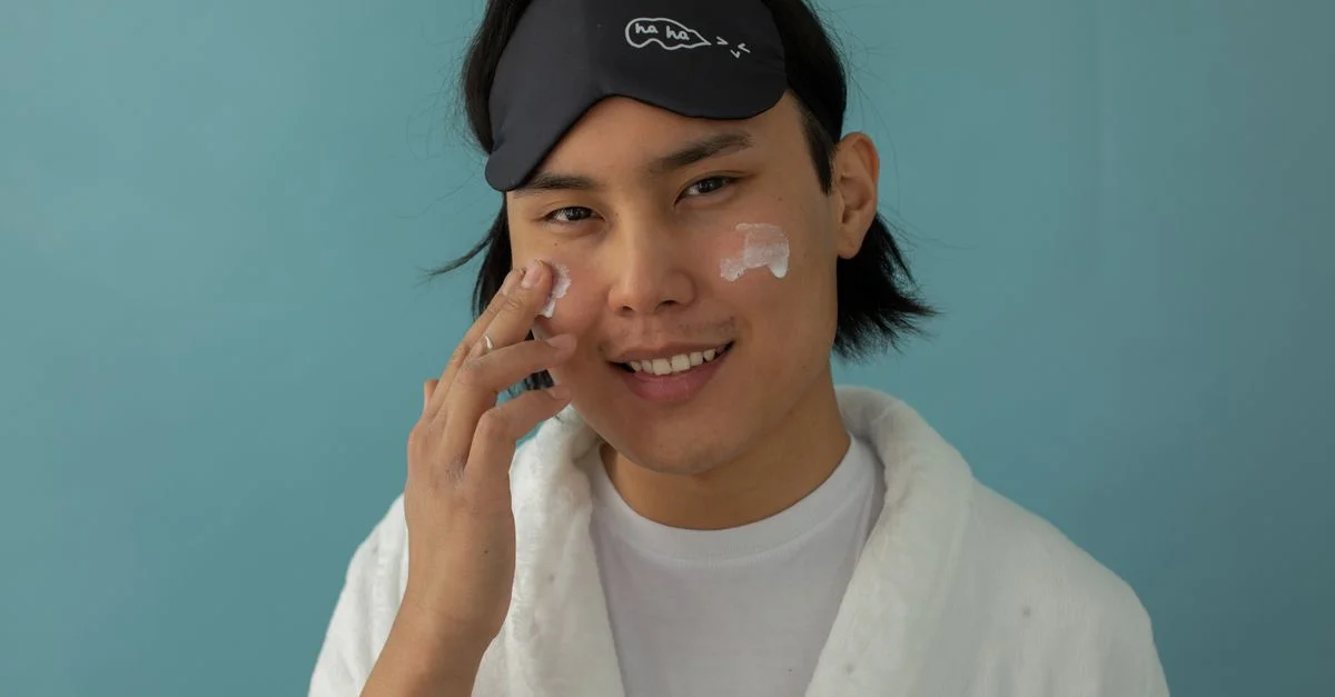 Hombre sonriente en bata aplicando crema facial con una máscara de sueño en un fondo azul.