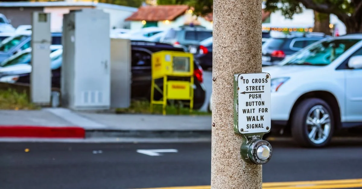 Botón de cruce peatonal en una calle urbana concurrida con coches estacionados cerca.
