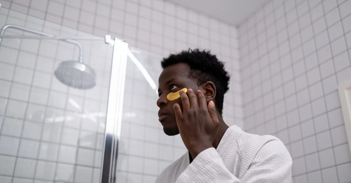 Hombre afroamericano con bata blanca aplicando parches para ojos en su rutina de cuidado de la piel en un baño contemporáneo.