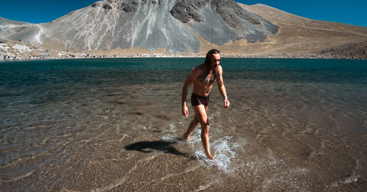 Un hombre camina por un sereno lago de montaña con agua azul clara y un impresionante telón de fondo rocoso.