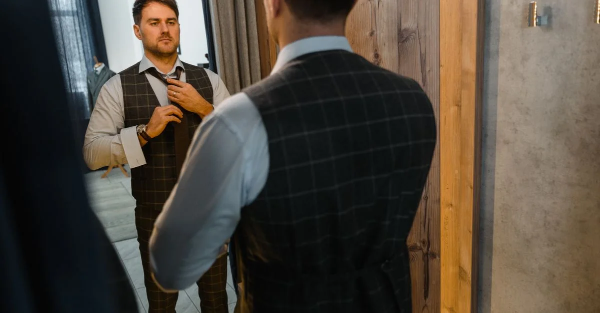 Hombre adulto con un chaleco elegante ajustando su corbata mientras se mira en el espejo en el interior.