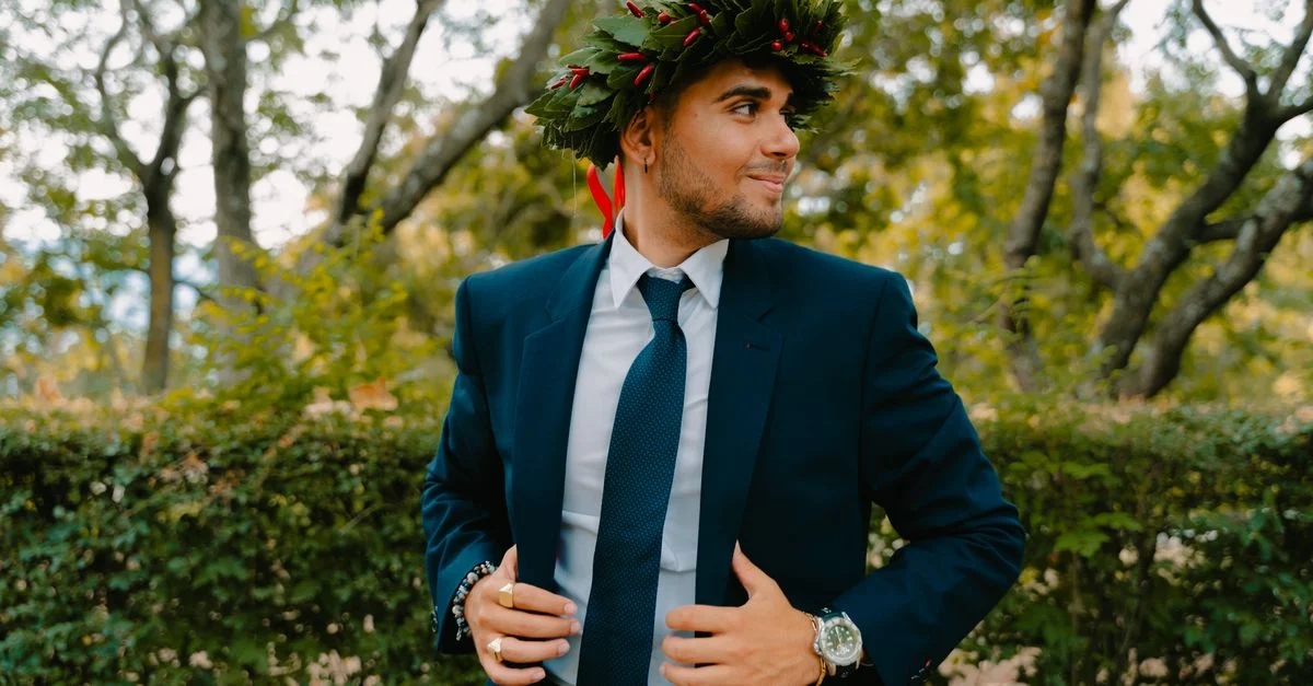 Joven en traje con una corona de laurel, sonriendo en un entorno exterior con hojas.